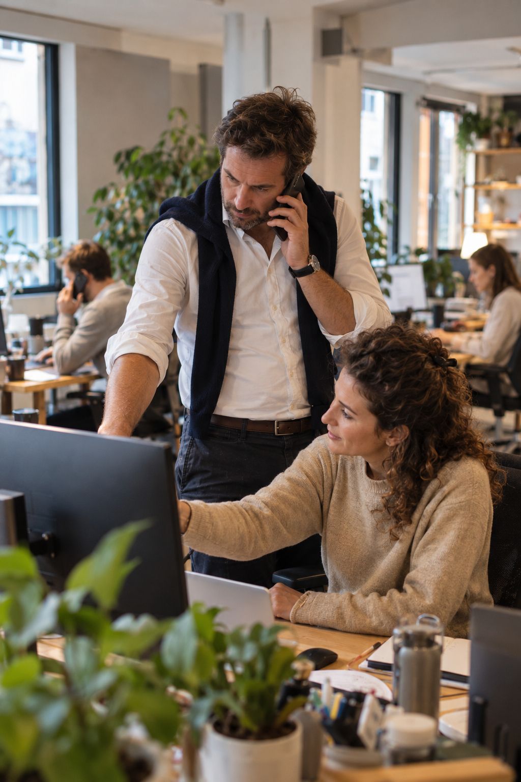 Dans les bureaux d’une PME, un responsable au téléphone se penche vers l’écran d’une collègue ; d’autres collègues travaillent en arrière-plan.